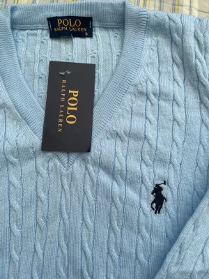 Ljusblå kabelstickad tröja Polo Ralph Lauren - Ljusblå kabelstickad tröja från Polo Ralph Lauren med V-ringning och klassisk broderad logga på bröstet. Tröjan har långa ärmar och ribbade muddar. Perfekt för dig som gillar stilrena och tidlösa plagg.