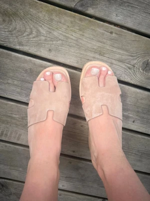 Beige slip-in platåsandaler i mocka - Snygga slip-in sandaler i beige mocka med breda band och utstansade detaljer. Sandalerna har en hög platåsula i kork-look som ger extra höjd och en trendig vibe. Perfekta till sommaren och lätta att matcha med olika outfits.