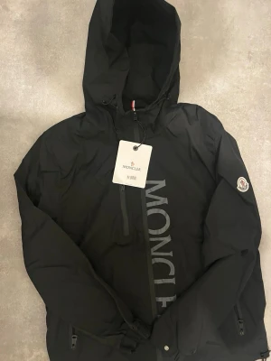 Svart vindjacka från Moncler - Snygg svart vindjacka från Moncler med stor logga längs framsidan och klassisk Moncler-patch på ärmen. Jackan har huva, bröstficka med dragkedja och två sidofickor. Tillverkad i lätt och slitstarkt material, perfekt för blåsiga dagar.