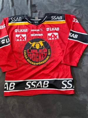 Luleå Hockey matchtröja signerad - Röd Luleå Hockey matchtröja från Bauer med klubbens stora logga på bröstet, svarta och gula detaljer samt flera sponsortryck. Tröjan har långa ärmar och är signerad av spelare. Perfekt för hockeyfans som vill sticka ut på läktaren.