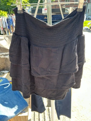 Svart volangkjol från BikBok - Supersöt svart kjol från BikBok med flera volanger och smock i midjan. Kjolen är kort och har en luftig känsla, perfekt för varma dagar. Enkel att matcha med olika toppar för en trendig look.