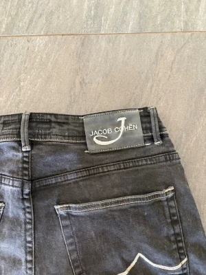 Svarta Jacob Cohën jeans - Snygga svarta jeans från Jacob Cohen med raka ben. Jeansen har kontrastsömmar på bakfickorna och en patch med logga bak i midjan. Tillverkade i mjukt denimtyg som ger en skön känsla och stilren look. Pris går att diskutera vid snabb affär 😊