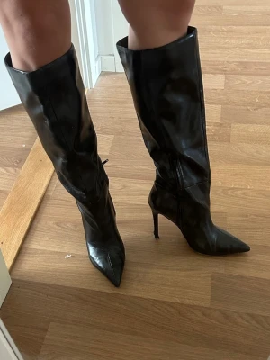 Svarta höga stövlar med klack - Snygga svarta stövlar i glansigt skinnimitation med spetsig tå och smal hög klack. Stövlarna går upp till knäna och har en stilren, trendig look som passar perfekt till kjol eller klänning.