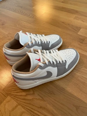 Air Jordan 1 Low grå/vit - Snygga Air Jordan 1 Low sneakers i vitt och grått läder med beige meshfoder. Klassisk röd Jumpman-logga på plösen och häl. Perforerad tåbox för extra ventilation och vita skosnören. Perfekta för dig som gillar stilrena och sportiga sneakers. Dom är andvända en gång. Säljs på grund av fel storlek 