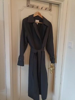 Mörkgrå lång trenchcoat med skärp - Stilren mörkgrå trenchcoat med klassisk krage, knytskärp i midjan och knappar framtill. Jackan har långa ärmar med dekorativa spännen vid ärmsluten och en rak, lång siluett. Perfekt för dig som gillar minimalistisk och tidlös stil.
