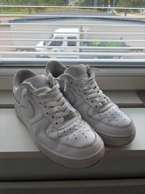 Vita Nike Air Force 1 sneakers - Klassiska vita Nike Air Force 1 sneakers i lågt utförande. Skorna har en ikonisk siluett med perforerad tå, vita skosnören och tydliga Nike-loggor på sidorna. Tillverkade i läder med gummisula för bra grepp. Perfekta för en clean och stilren look.
