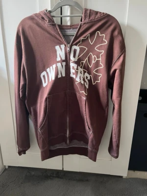 Brun hoodie med tryck från No Owners - Säljer en brun hoodie med dragkedja från No Owners. Framsidan har vitt tryck med texten 'NO OWNERS' och ett stort löv i beige. På huvan finns ett mindre lövtryck. Hoodien har fickor fram och ribbade muddar vid ärmslut och nederkant. Perfekt för en avslappnad stil.