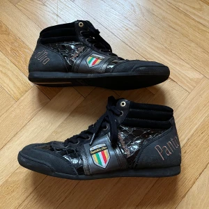 Svarta sneakers från Pantofola d'Oro - Snygga svarta sneakers från Pantofola d'Oro med krokodilmönstrade detaljer och färgglada emblem på sidan. Skorna har snörning och en högre ankel för extra stöd. Perfekta för en stilren look med en touch av unik design.