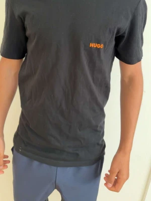 Svart t-shirt från HUGO - Säljer en svart t-shirt från HUGO med orange logga på bröstet. Klassisk passform och rund halsringning, perfekt till jeans eller shorts. Enkel och stilren design som funkar till det mesta.