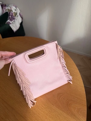 Rosa maje väska  - Säljer en ljusrosa handväska från Maje med rektangulärt handtag och coola fransdetaljer på sidorna. Väskan är gjord i ett strukturerat material som ger en trendig look. Perfekt accessoar för att lyfta din outfit. Väldigt gott skick då jag knappt andvänd den. Stora storleken. Långa bandet medföljer 