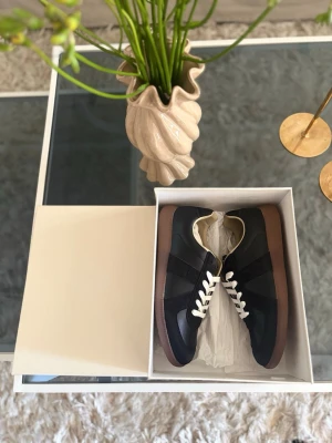 Maison Margiela - Snygga svarta sneakers med vita skosnören och brun gummisula. Ovandelen är i en mix av mocka och slätt läder, vilket ger en stilren och klassisk look. 