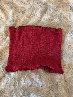 Röd smockad tubtopp - Trendig röd tubtopp med smockad struktur och vågiga kanter upptill och nedtill. Perfekt att styla till jeans eller kjol för en cool och enkel look. Materialet är stretchigt och följsamt för en bekväm passform.