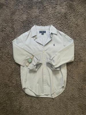 Ljusblå skjorta från Ralph Lauren - Klassisk ljusblå skjorta från Ralph Lauren med broderad logga på bröstet. Skjortan har lång ärm, knappar framtill och en avslappnad passform. Perfekt till jeans eller chinos för en clean och stilren look.