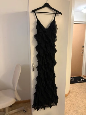 Svart volangklänning med smala band - Lång svart klänning med smala axelband och massor av volanger längs hela klänningen. Snygg och dramatisk look med omlottkänsla och v-ringad hals. Perfekt för dig som vill sticka ut lite extra.