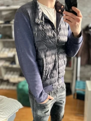 Mörkblå cardigan från Ralph lauren - Snygg mörkblå pufferjacka från Ralph lauren med quiltad kropp och stickade ärmar. Jackan har dragkedja framtill, två fickor med tryckknappar och ribbade muddar vid ärmslut och nederkant. Perfekt för dig som vill ha en sportig och trendig look.