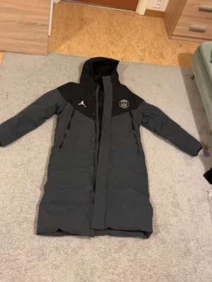 Svart/grå lång dunjacka PSG  - Lång och varm dunjacka i svart och mörkgrått från PSG. Jackan har huva, dragkedja framtill, två sidofickor med dragkedja och PSG-logga samt Jumpman-logga på bröstet. Perfekt för kalla vinterdagar och riktigt snygg streetstil.