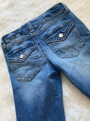 Blå jeans från Gina Tricot - Säljer ett par klassiska blå jeans från Gina Tricot med snygga bakfickor med lock och silverfärgade knappar. Jeansen har normal passform och är gjorda i mjuk bomull med lite stretch. Perfekta till alla dina outfits!