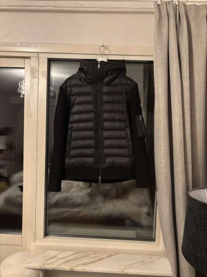 Svart cardigan med luva från Moncler - Snygg svart cardigan från Moncler med huva och dragkedja framtill. Västen har stickade ärmar och en liten ficka med dragkedja på ena ärmen samt Moncler-logga. Perfekt för kyliga dagar och riktigt stilren look.