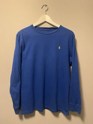 Blå långärmad tröja från Polo Ralph Lauren - Snygg blå långärmad tröja från Polo Ralph Lauren med klassisk gul broderad logga på bröstet. Tröjan har rund halsringning och är gjord i mjuk bomull för en skön känsla. Perfekt för dig som gillar stilrena och tidlösa plagg. Kostar runt 1000 lappen ny!