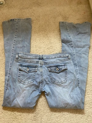 Ljusblå bootcut jeans - ljusblå bootcut jeans i klassisk denim med ficklock och knappdetaljer bak. Jeansen har låg midja och utsvängda ben, perfekta för en avslappnad och trendig look. Materialet är mjukt och följsamt bomullsdenim.