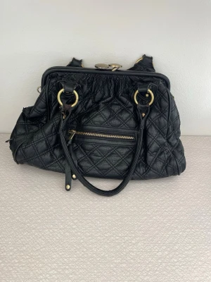Marc Jacobs svart quiltad handväska - Cool svart handväska från Marc Jacobs i quiltat läder med guldfärgade metalldetaljer och dragkedja framtill. Väskan har dubbla handtag och snygg knäppning med två runda knappar upptill. Perfekt för dig som gillar stilrena och trendiga accessoarer.