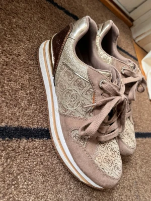 Beige och guldiga sneakers från Guess - Snygga sneakers från Guess i beige och guld med logomönster över hela skon. Skorna har glittriga detaljer, metallicbrun häl och vita sulor med guldinslag. Klassisk snörning och Guess-logga på sidan och sulan. Perfekta för dig som gillar en lyxig och trendig look.