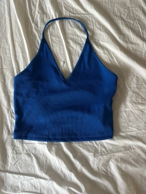 Blå ribbad halterneck topp - Snygg blå ribbad linnetopp med halterneck och v-ringning. Toppen är croppad och har en enkel, stilren design som passar perfekt till jeans eller kjol. Materialet känns mjukt och stretchigt för en bekväm passform.
