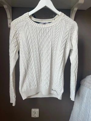Vit kabelstickad tröja från Bondelid  - Kabelstickad vit tröja från Bondelid med rund halsringning och ribbade muddar. Tröjan har ett broderat Bondelid-märke vid nederkanten och en patch på ena ärmen. 