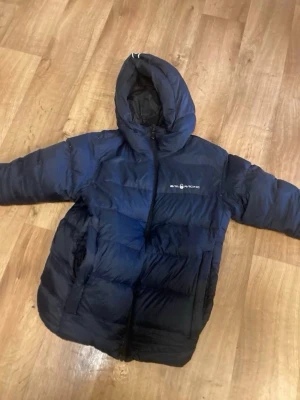 Mörkblå dunjacka med huva - Snygg mörkblå dunjacka från Everest med huva och dragkedja framtill. Jackan har en puffig design och logga på bröstet. Perfekt för kalla vinterdagar när du vill hålla dig varm och bekväm.