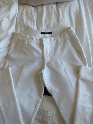Vita kostymbyxor från Bik Bok - Stilrena vita kostymbyxor från Bik Bok med klassisk passform och pressveck. Byxorna har fickor i sidorna och dold dragkedja framtill. Perfekta för en dressad look och enkla att matcha med olika toppar. Liten i storleken så passar storlek 36 bäst. 