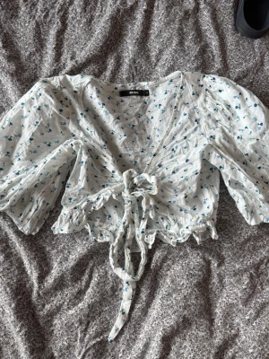 Blommig knytblus från Bik Bok - Supersöt blus från Bik Bok i vitt med blått blommönster. Modellen har puffiga ärmar, djup v-ringning och knyt framtill. Volangkant nedtill ger en extra feminin touch. Perfekt för dig som gillar romantisk och trendig stil.