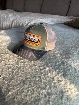 Stetson truckerkeps grön och beige - Snygg truckerkeps från Stetson med grön front, beige mesh baktill och svart böjd skärm. Broderad patch framtill med texten 'Stetson American Heritage' i orange och vitt. Perfekt accessoar för en avslappnad streetstil.