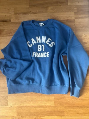 Blå sweatshirt med Cannes-tryck - Blå sweatshirt med texten 'CANNES 91 FRANCE' i vitt på bröstet. Tröjan har rund halsringning, ribbade muddar vid ärmslut och nederkant. Perfekt för en avslappnad och sportig stil. Materialet känns mjukt och bekvämt, troligen bomullsblandning.