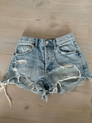 Ljusa slitna jeansshorts med fransar - Säljer ett par ljusblå denimshorts med hög midja och råa, fransiga kanter. Shortsen har slitna detaljer framtill, klassiska fem fickor och bälteshällor. Perfekta för sommaren och ger en avslappnad, trendig look.