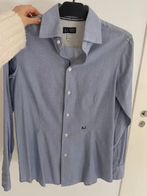 Blå randig skjorta från Armani Jeans - Klassisk blå skjorta från Armani Jeans med diskreta vita ränder och broderad AJ-logga på bröstet. Skjortan har lång ärm, vit knappstängning och en tidlös krage. Perfekt för en clean och stilren look.