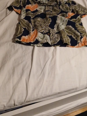 Blommig skjorta från Lindex - Kortärmad skjorta från Lindex med stort blommigt mönster i grönt, orange och vitt på mörkblå botten. Skjortan har klassisk krage och knappar framtill, perfekt för en avslappnad och trendig look.