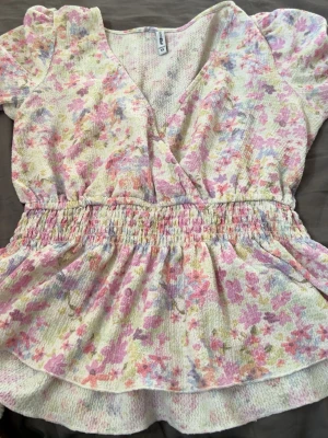 Blommig omlottopp från H&M - Söt blommig omlottopp från H&M med korta puffärmar och smockad midja. Toppen har ett ljust mönster i rosa, lila, gul och grön på vit botten. Tillverkad i ett mjukt, strukturerat material som känns bekvämt mot huden. Perfekt för vår och sommar! Stor i storlek då den passar mig som XL