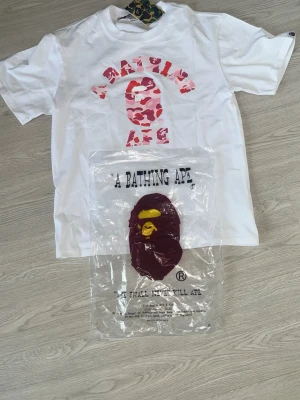 Vit BAPE t-shirt med rosa camo - Säljer en clean vit t-shirt från A Bathing Ape med det klassiska BAPE-huvudet i rosa camo och röd text på bröstet. T-shirten har korta ärmar och är gjord i mjuk bomull. Perfekt för dig som gillar streetwear och vill sticka ut lite extra.