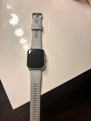 Grå smartklocka med silikonarmband - Stilren smartklocka med fyrkantig display och grått silikonarmband. Klockan har silverfärgad ram och flera justeringshål för perfekt passform. Baksidan har sensorer för pulsmätning och laddning. Perfekt för dig som vill ha koll på hälsa och notiser direkt på handleden. Funkar både android och apple.