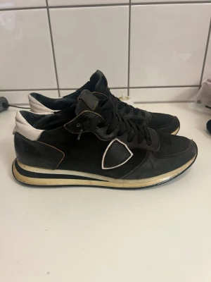 Svarta sneakers från Philippe Model - Säljer ett par svarta Philippe Model sneakers med vita detaljer och logga på sidan. Skorna har svart mesh och läder, vita hälpartier och en vit sula med svarta och beige inslag. Klassisk låg modell med snörning och sportig look.