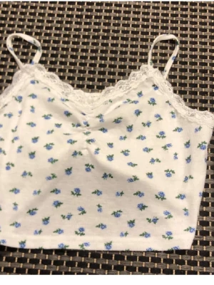 Vit bralette med blå blommor och spets - Söt vit bralette med tunna axelband och spetskant upptill. Tyg i mjuk bomull med små blå blommor över hela plagget. Ingen bygel och utan vaddering för en bekväm känsla. Perfekt för dig som gillar romantisk och enkel stil.
