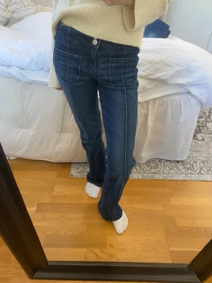 Mörkblå raka jeans med stora fickor - Säljer ett par mörkblå jeans med raka ben och stora, markerade framfickor. Jeansen har tydliga sömmar framtill och klassisk knappgylf. Perfekta för en avslappnad och trendig look.