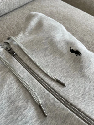 Grå zip hoodie från Polo Ralph Lauren - Snygg ljusgrå hoodie med dragkedja från Polo Ralph Lauren. Klassisk design med huva och dragsnören, samt den ikoniska svarta loggan broderad på bröstet. Tillverkad i mjukt bomullsmaterial som är skönt att bära. Perfekt till en avslappnad stil. Använd sparsamt och inga tecken på fläckar eller defekter osv. Skick 9/10. Om du har några funderingar, tveka inte på att skriv. Kan gå ner i pris vid snabb affär. Passa på!