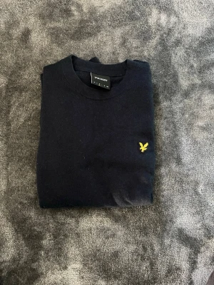 Mörkblå tröja från Lyle & Scott - Snygg mörkblå tröja från Lyle & Scott med klassisk rund hals och den ikoniska gula örnloggan broderad på bröstet. Tröjan har långa ärmar och är tillverkad i mjukt material som känns skönt mot huden. Perfekt för dig som gillar stilrena och tidlösa plagg.