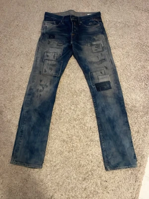 Unika Replay Jeans - Unika blå jeans med raka ben och coola patchade detaljer framtill. Jeansen har en tvättad look och flera dekorativa lappar i olika storlekar som ger en trendig och avslappnad stil. Perfekta för dig som vill sticka ut med din outfit.
