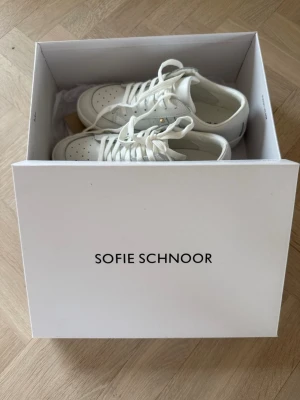 Vita sneakers från Sofie Schnoor - Stilrena vita sneakers från Sofie Schnoor i läder med klassisk låg profil och perforerade detaljer på tån. Skorna har vita snören och subtila gulddetaljer på sidan. Perfekta för en clean och modern look.