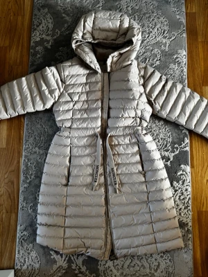 Ljusbeige lång dunjacka - Snygg ljusbeige lång dunjacka med huva och dragkedja framtill. Jackan har ett bälte i midjan och quiltad design. Perfekt för kalla vinterdagar och riktigt varm tack vare dunfyllningen.