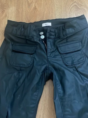 Svarta coated jeans från Nelly - Säljer ett par svarta coated jeans från Nelly med två stora fickor framtill och klassiska bakfickor. Jeansen har låg midja, utsvängda ven och dubbla silverknappar i linningen. Materialet har en glansig läder liknande finish som ger en cool look. Dem är avklippta längst ner men bra längd för mig som är 160. 