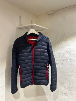 Mörkblå pufferjacka med röda detaljer - Snygg mörkblå pufferjacka från Cross Sportswear med röda dragkedjor och detaljer. Jackan har ståkrage, två framfickor med dragkedja och en liten röd logga på bröstet. Perfekt för kalla dagar och ger en sportig look.