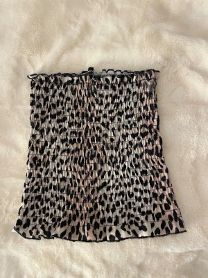 Leopardmönstrad tubtopp i beige och svart - Trendig tubtopp med leopardmönster i beige och svart. Smockad och stretchig modell som sitter snyggt på kroppen. Perfekt att styla till jeans eller kjol för en cool look.